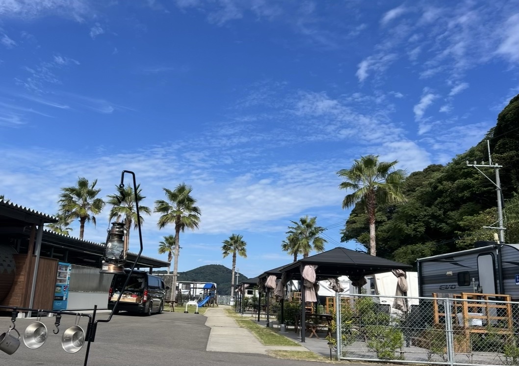 ヤシの木が並ぶ、呼子のトレーラーハウスビレッジのRVキャンプエリア。青空の下、ランタンとハイエースが見える
